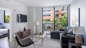 Ein modernes Wohnzimmer mit Sofa, Sessel, Couchtisch, Fernseher und einem Balkon mit Blick auf Häuser und Grünflächen.