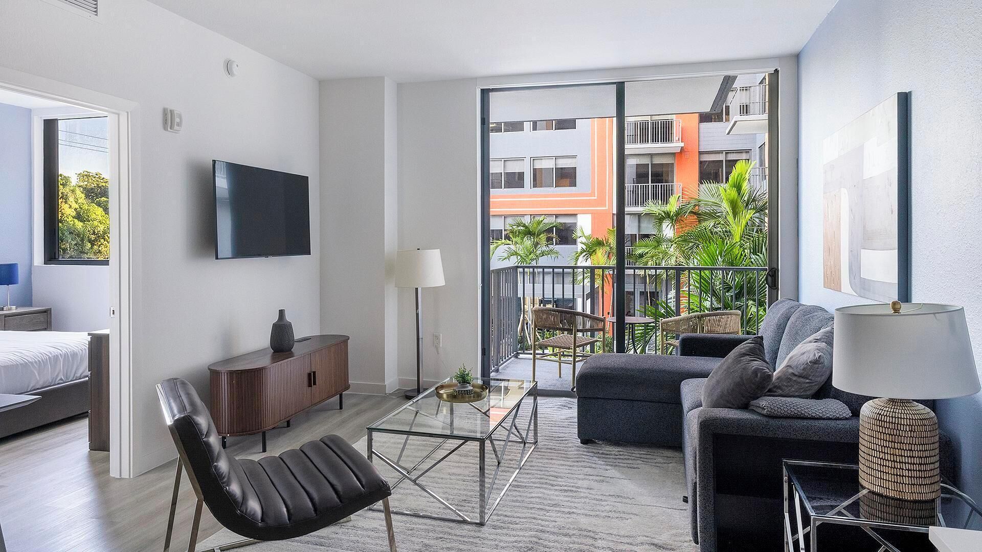 A modern living room with a sofa, armchair, coffee table, TV, and a balcony with a view of buildings and greenery.
