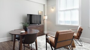 Ein modernes Wohnzimmer mit einem Flachbildfernseher, einem Esstisch mit Stühlen, einer Zimmerpflanze und einem großen Fenster mit Jalousien.