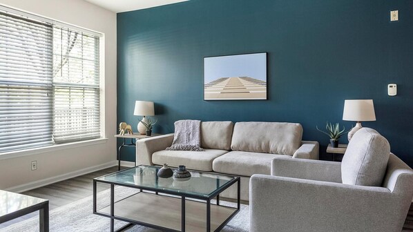 A living room with a blue accent wall, a sofa, a glass coffee table, and two lamps. - Landing | Incredible 2BD, Gym, Pool (King of Prussia)