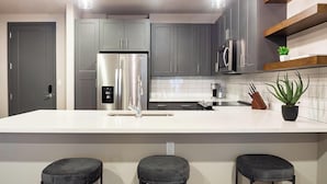 A modern kitchen with grey cabinets, a stainless steel refrigerator, a microwave, a sink, and a bar stool area. - Landing | Spectacular 1BD, Clubhouse (Tulsa)
