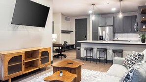 A modern living room with a flat-screen TV, wooden furniture, a kitchen with grey cabinets, and a bar area. - Landing | Stylish 1BD, Clubhouse (Tulsa)