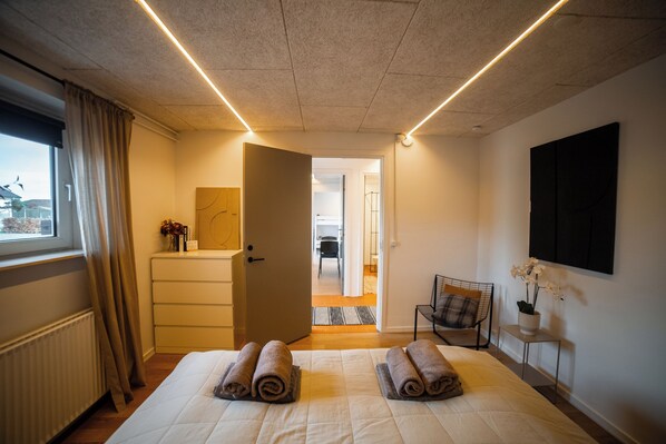 A hotel room with a bed, a dresser, a chair, a small table with a plant, and a window with curtains.