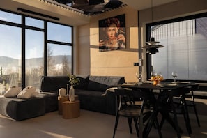 A modern living room with a sofa, dining table, and chairs, large windows with a view of mountains, and a painting on the wall. - Flowfort View Private Hottub & Sauna Dom Swobodnie (Stara Kamienica)