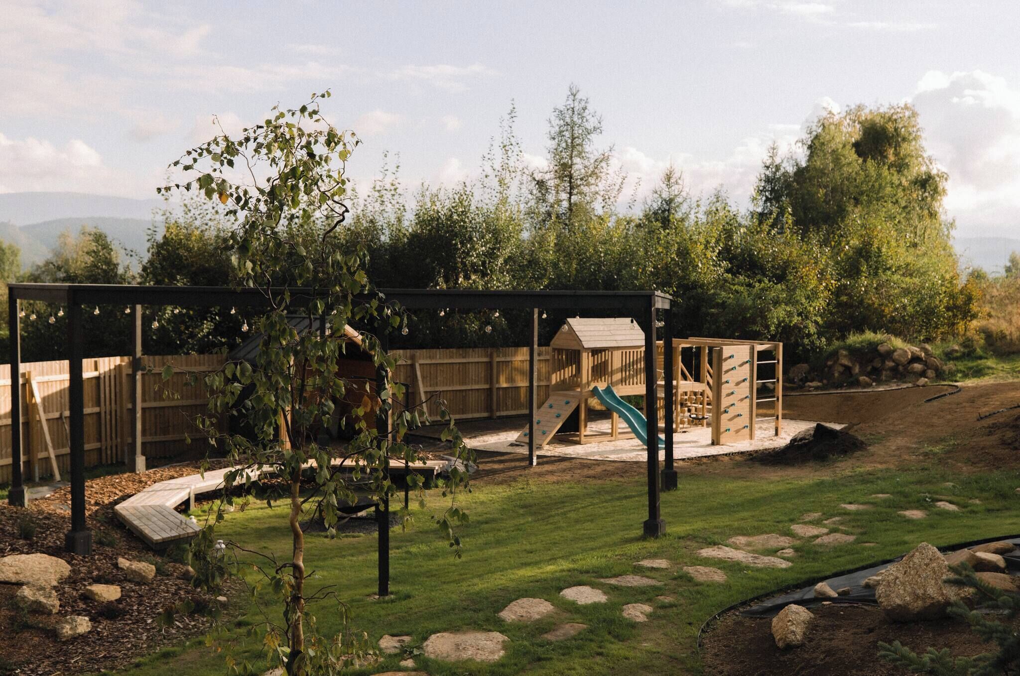 Un parque infantil con tobogán y estructuras de escalada, rodeado por una valla de madera y árboles.