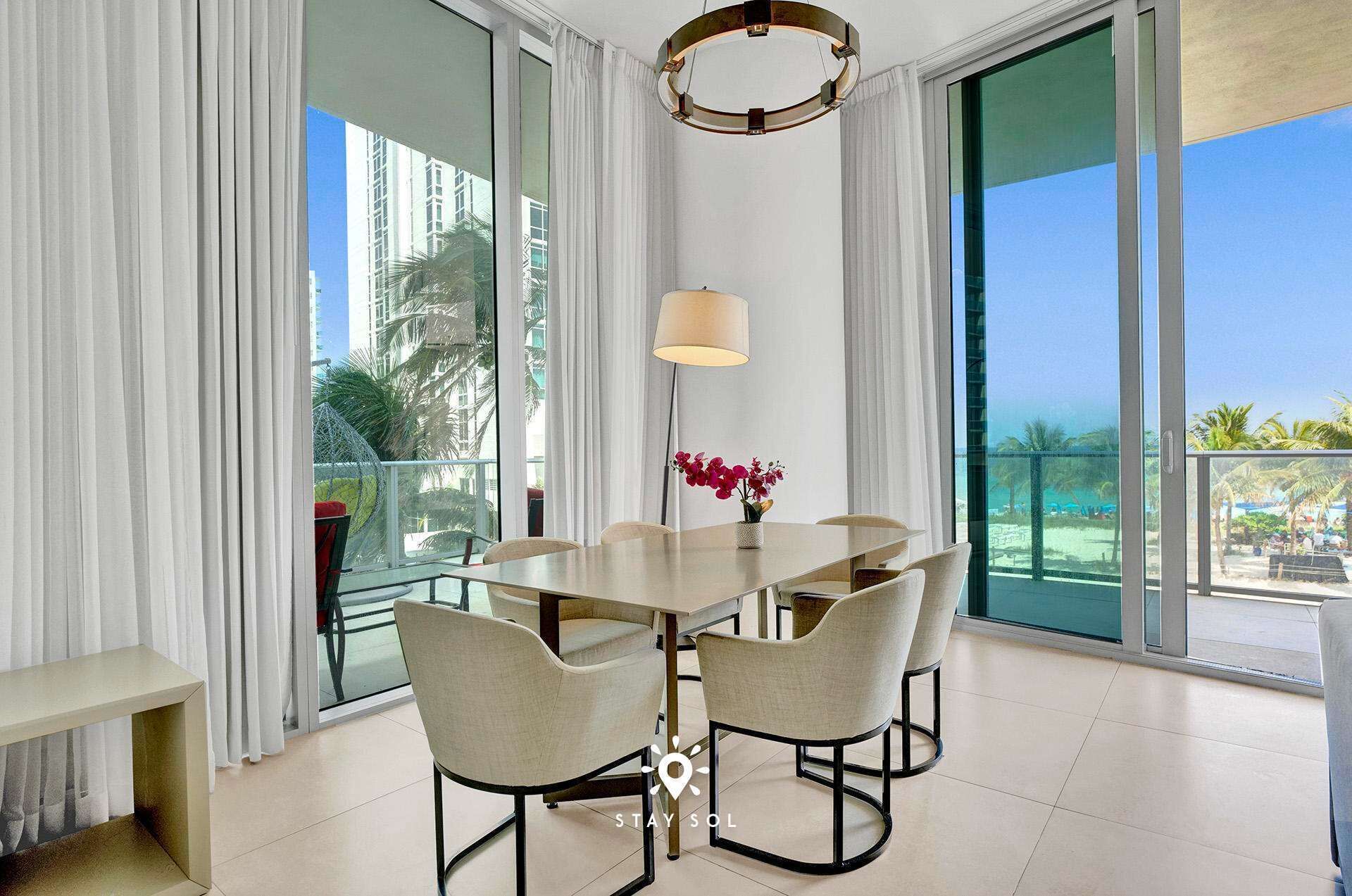 Un espace repas moderne avec une table ronde, six chaises, un vase rempli de fleurs rouges et une grande fenêtre donnant sur la plage et les palmiers.
