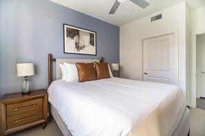 Une chambre à coucher comprenant un lit, une table de chevet, une lampe, un ventilateur de plafond, une porte et un tableau encadré accroché au mur.