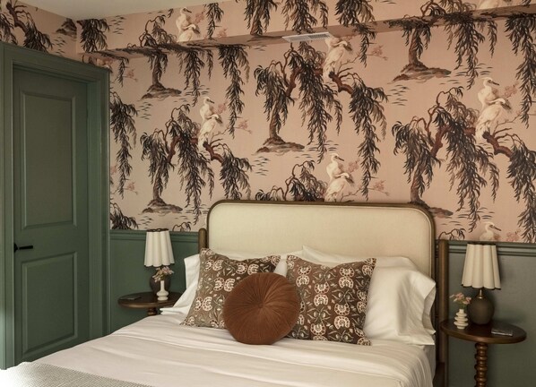 Une chambre à coucher avec un lit, deux tables de chevet, une commode, une porte verte et un mur recouvert d’un papier peint à motifs naturels.
