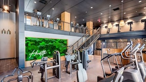Un gimnasio moderno con varios equipos de ejercicio, una pared verde con la palabra 'Hustle' y una escalera que conduce a un nivel superior.