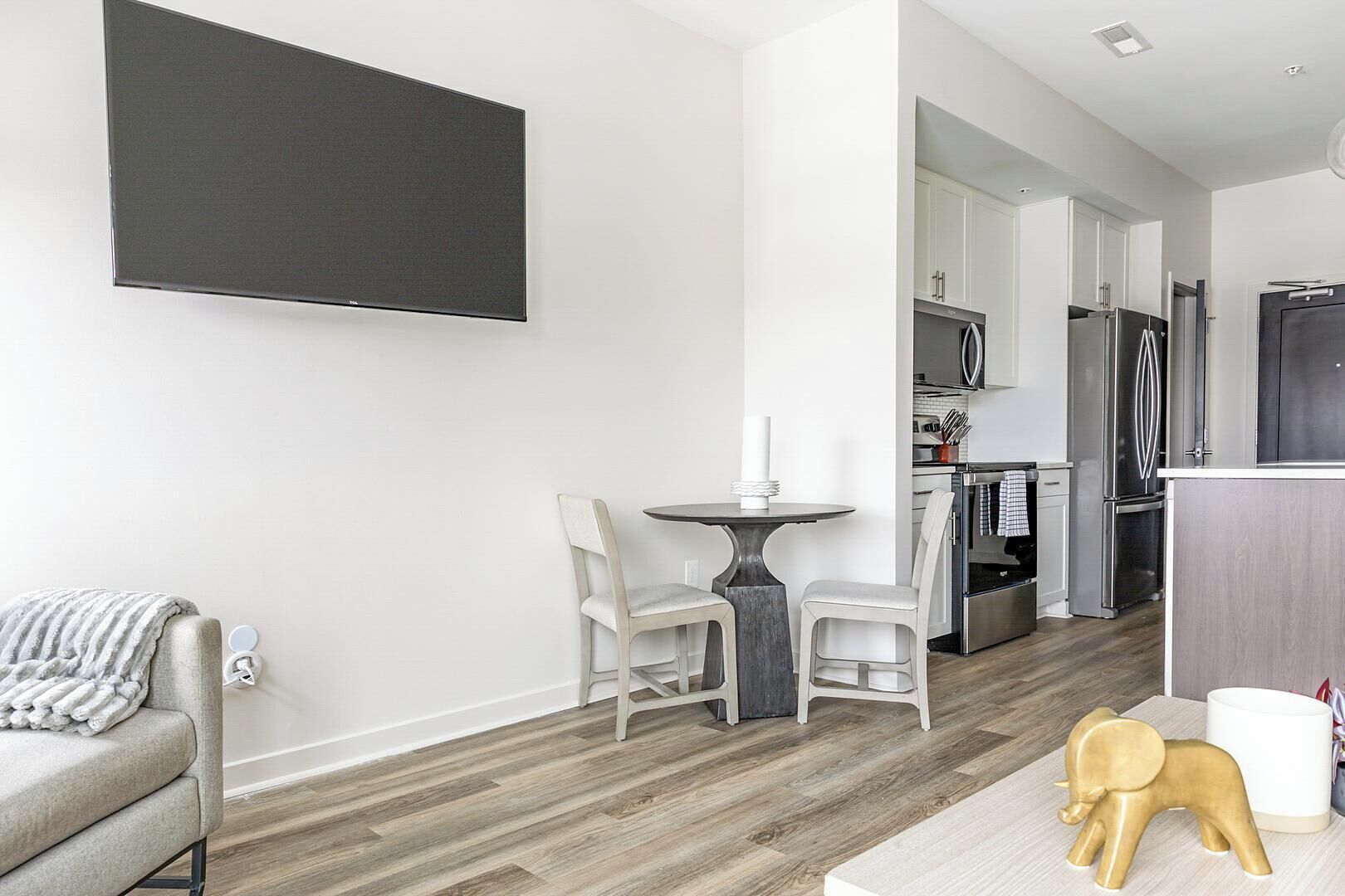 Ett modernt vardagsrum med en grå soffa, ett matbord med två stolar, en TV som är monterad på väggen och ett köksområde med vitvaror i rostfritt stål.