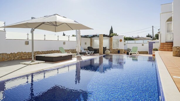 An indoor swimming pool with lounge chairs and an umbrella, surrounded by stone walls and a clear sky. - Casa Descoberta - Luxurious 4 Bedroom family Villa with heated pool (Carvoeiro)