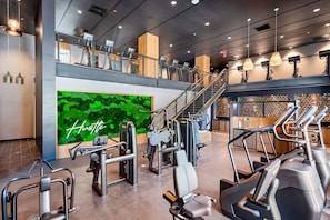 A modern gym with various exercise equipment, a green wall with the word 'Hustle,' and a staircase leading to an upper level. - Landing | Modern 1BD, Yoga Studio, Clubhouse (Columbus)