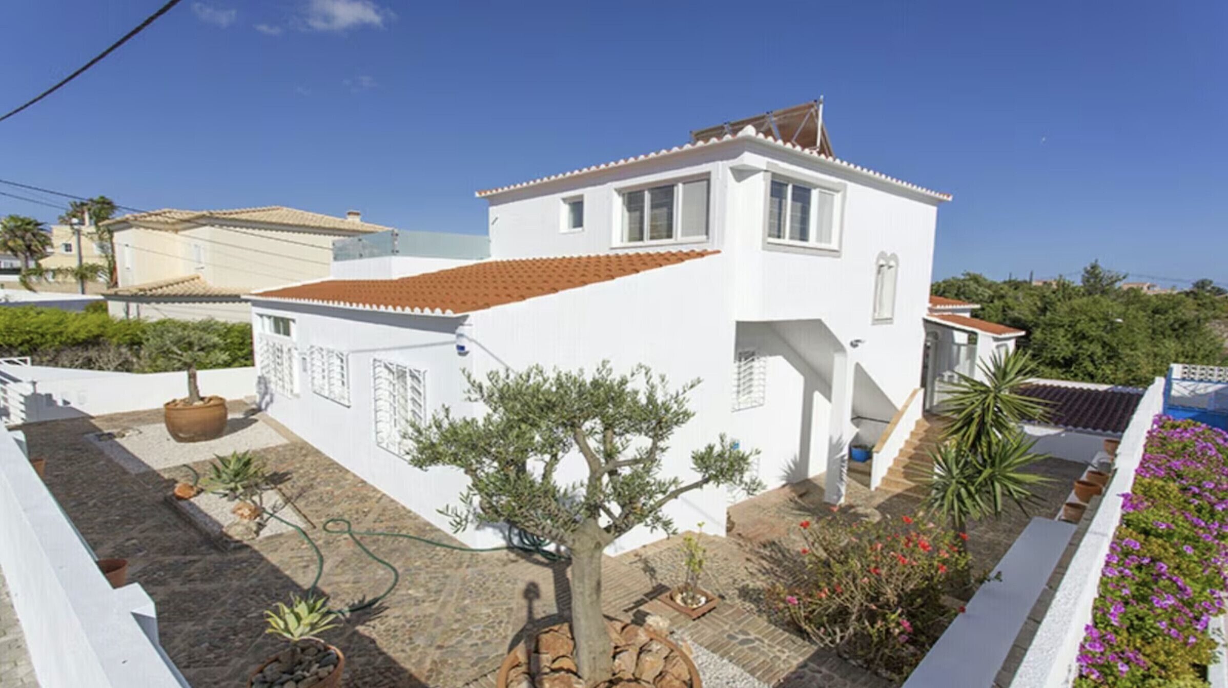 A white two-story house with a tiled roof, a small garden with potted plants, and a staircase leading to the entrance.