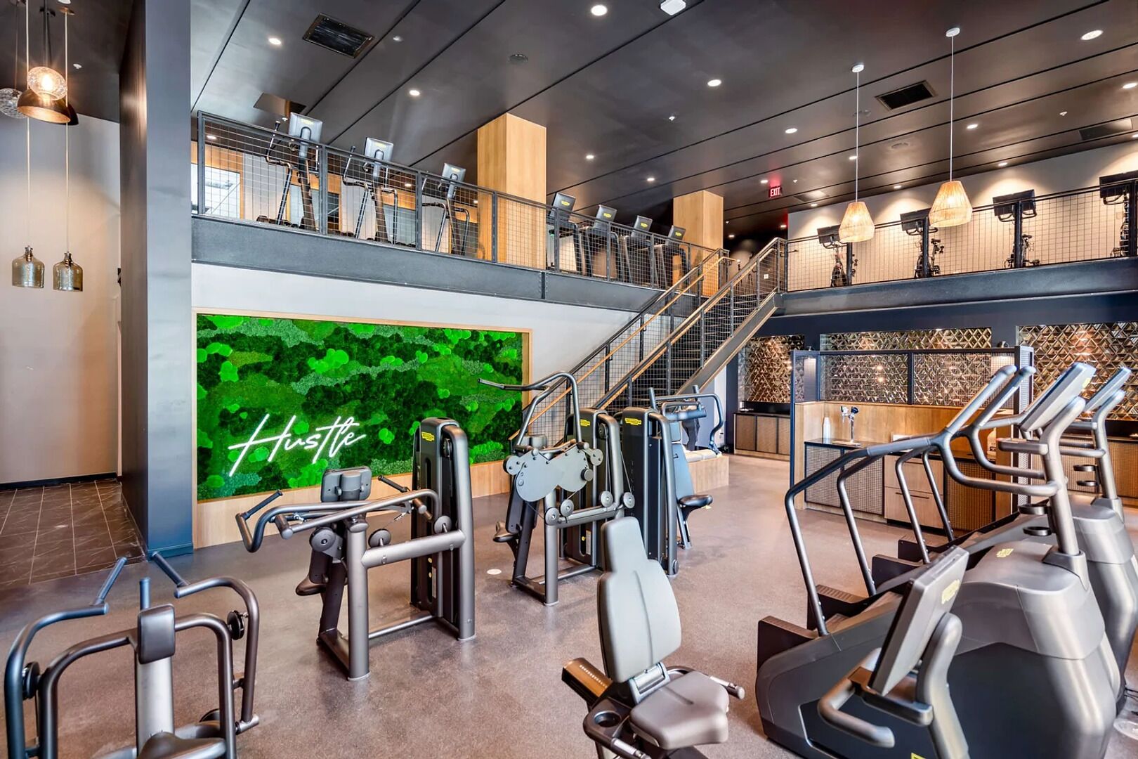 Una palestra moderna con diverse attrezzature per l'allenamento, un muro verde con la scritta 'Hustle' e una scala che conduce a un piano superiore.