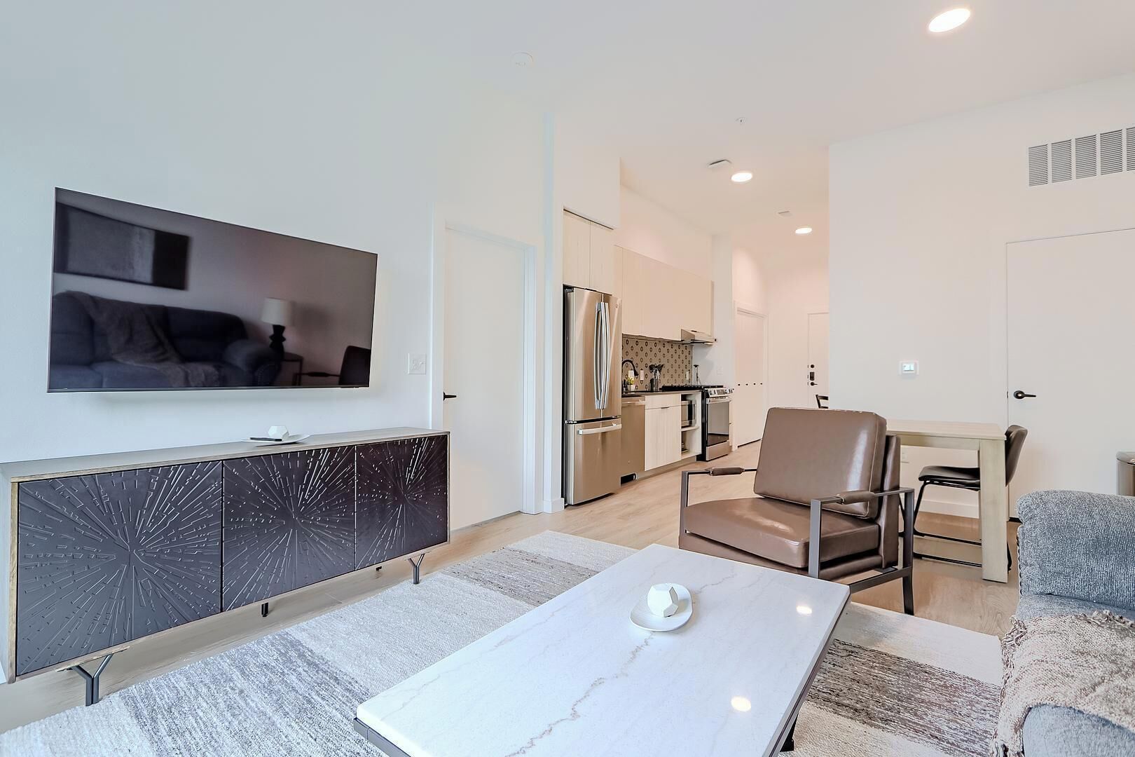 A modern living room with a flat-screen TV, a sofa, a dining table, and a kitchen in the background.