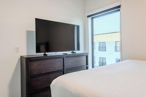 Een slaapkamer met een bed, een kaptafel, een televisie en een raam met uitzicht op gebouwen.