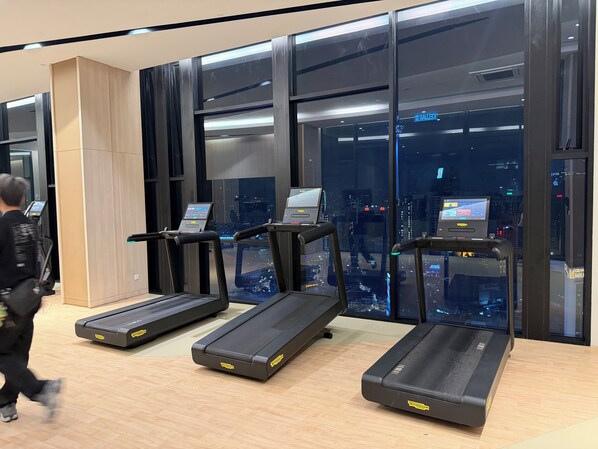 Ein Fitnessraum mit Laufbändern, einer Bedienkonsole und Blick auf die Stadt durch große Fenster.