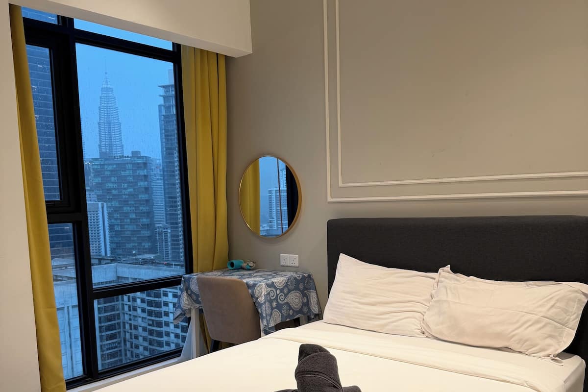 Ein modernes Hotelzimmer mit einem großen Bett, einem runden Spiegel, Blick auf eine Stadtlandschaft durch ein großes Fenster und einem kleinen Tisch mit Stuhl.
