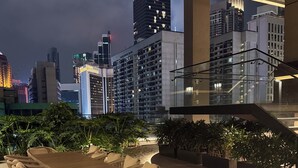 Uma área de refeições ao ar livre com mesa e cadeiras, rodeada por plantas em vasos, com vista para um horizonte urbano com edifícios altos.