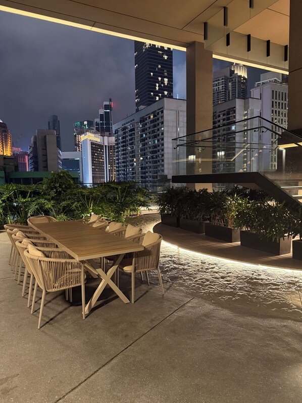 Un espace extérieur aménagé pour les repas, avec une table et des chaises, entouré de plantes en pot, et offrant une vue sur un paysage urbain avec de hauts immeubles.