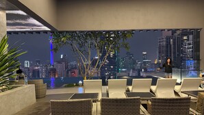 Um lounge no terraço com cadeiras de vime, vista para a cidade à noite e uma pessoa falando.