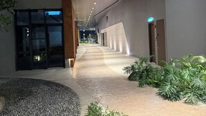 Um corredor com teto moderno, entrada por porta de vidro e um caminho ladeado por plantas.