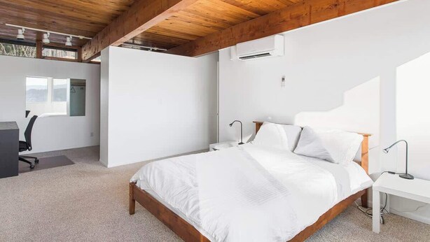 A bedroom with a wooden ceiling, a bed with white bedding, a desk with a chair, and a window.