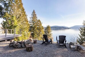 Una terraza junto al lago con sillas Adirondack, un pozo de fuego y una cubierta de madera con vistas a un lago tranquilo.