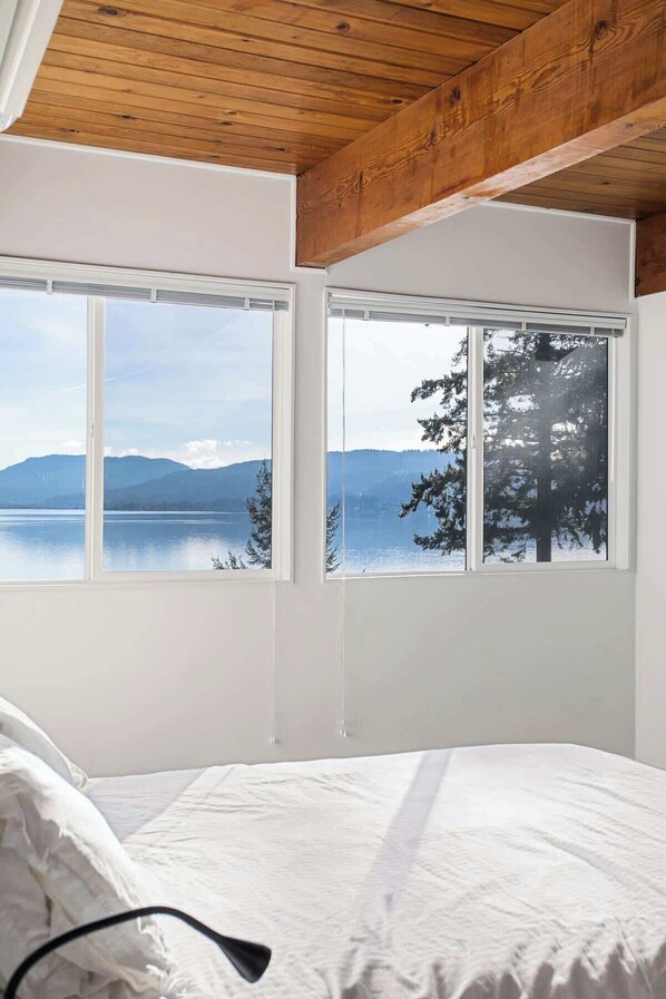 Un dormitorio con un ventanal grande que da a un lago y montañas, techo de madera y cama blanca.