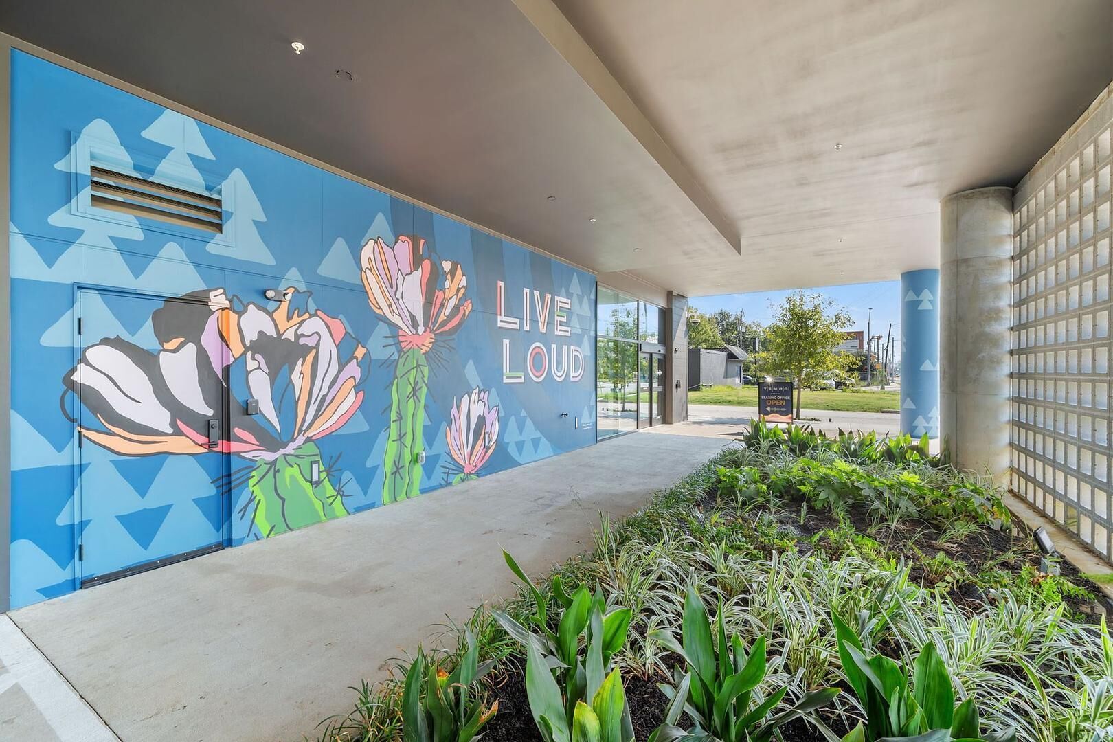 A building exterior with a mural, a sign reading 'LIVE LOUJD,' and a landscaped area with plants.