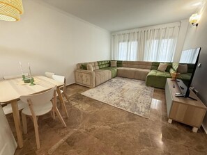 A modern living room with a sectional sofa, a dining table with chairs, a flat-screen TV, and a large window with curtains. - Beachside Apartment - Walk to the Sea, Minutes to Puerto Banús (San Pedro de Alcántara)