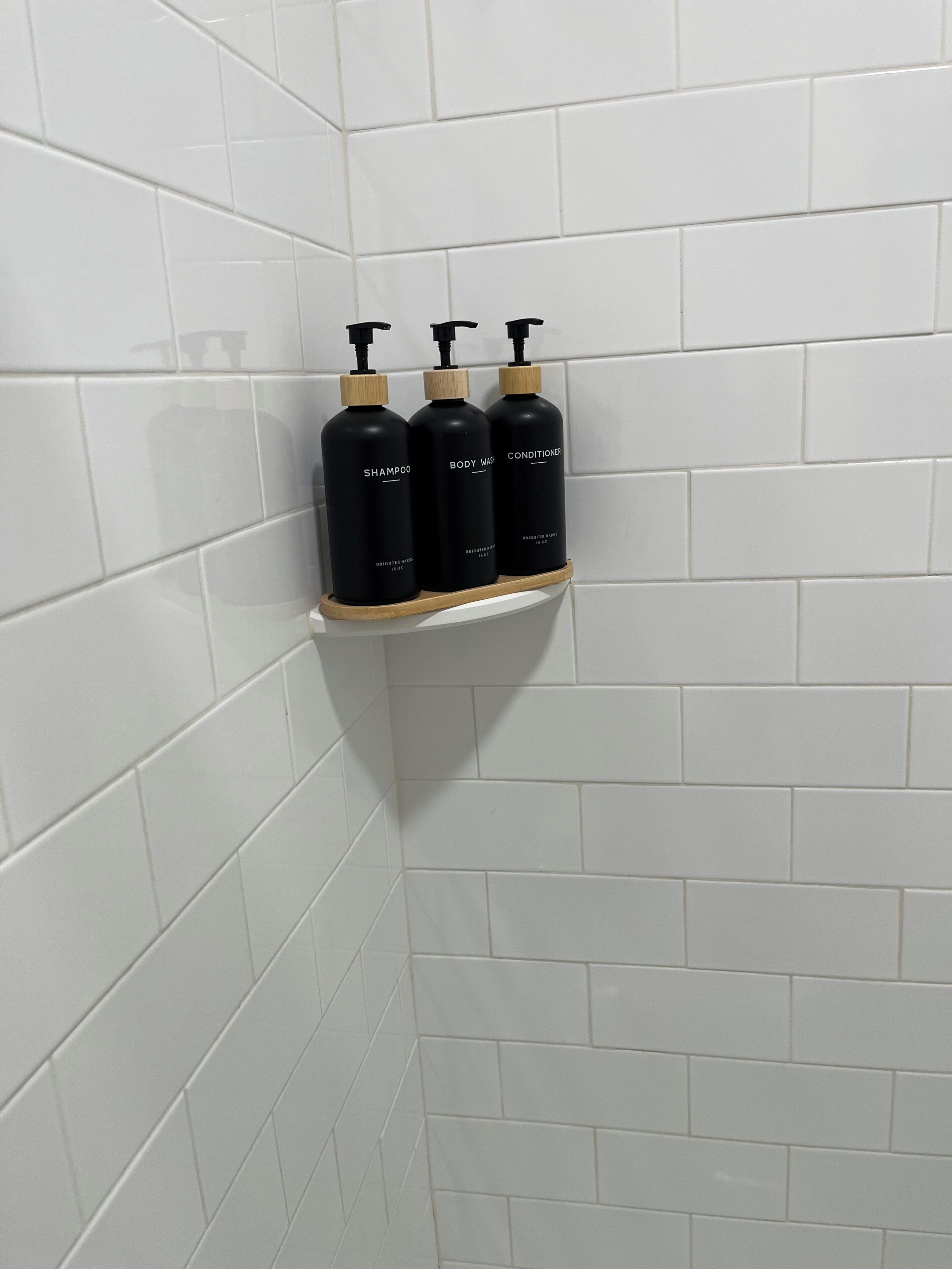 Un coin de salle de bain avec des carreaux blancs et une étagère sur laquelle reposent trois bouteilles noires étiquetées « SHAMPOO », « GEL DOUCHE » et « APRÈS-SHAMPOO ».