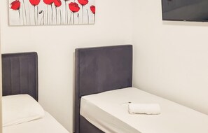 Deux lits simples avec des têtes de lit grises et une literie blanche, un téléviseur à écran plat fixé au mur et un tableau représentant des fleurs rouges au-dessus des lits.