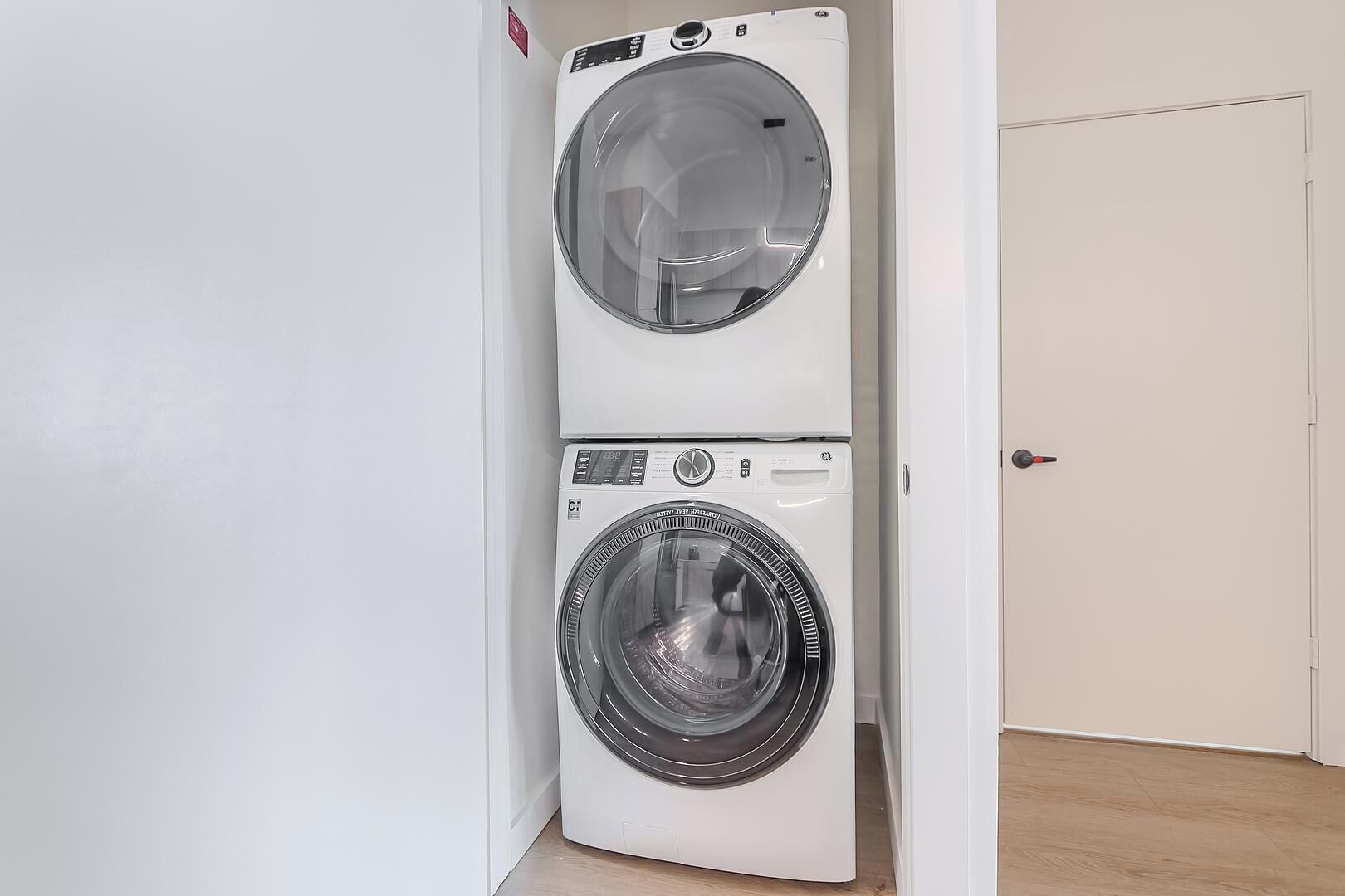 Uma máquina de lavar e secar compacta, branca e de parede, em um corredor com piso de madeira e uma porta fechada ao fundo.