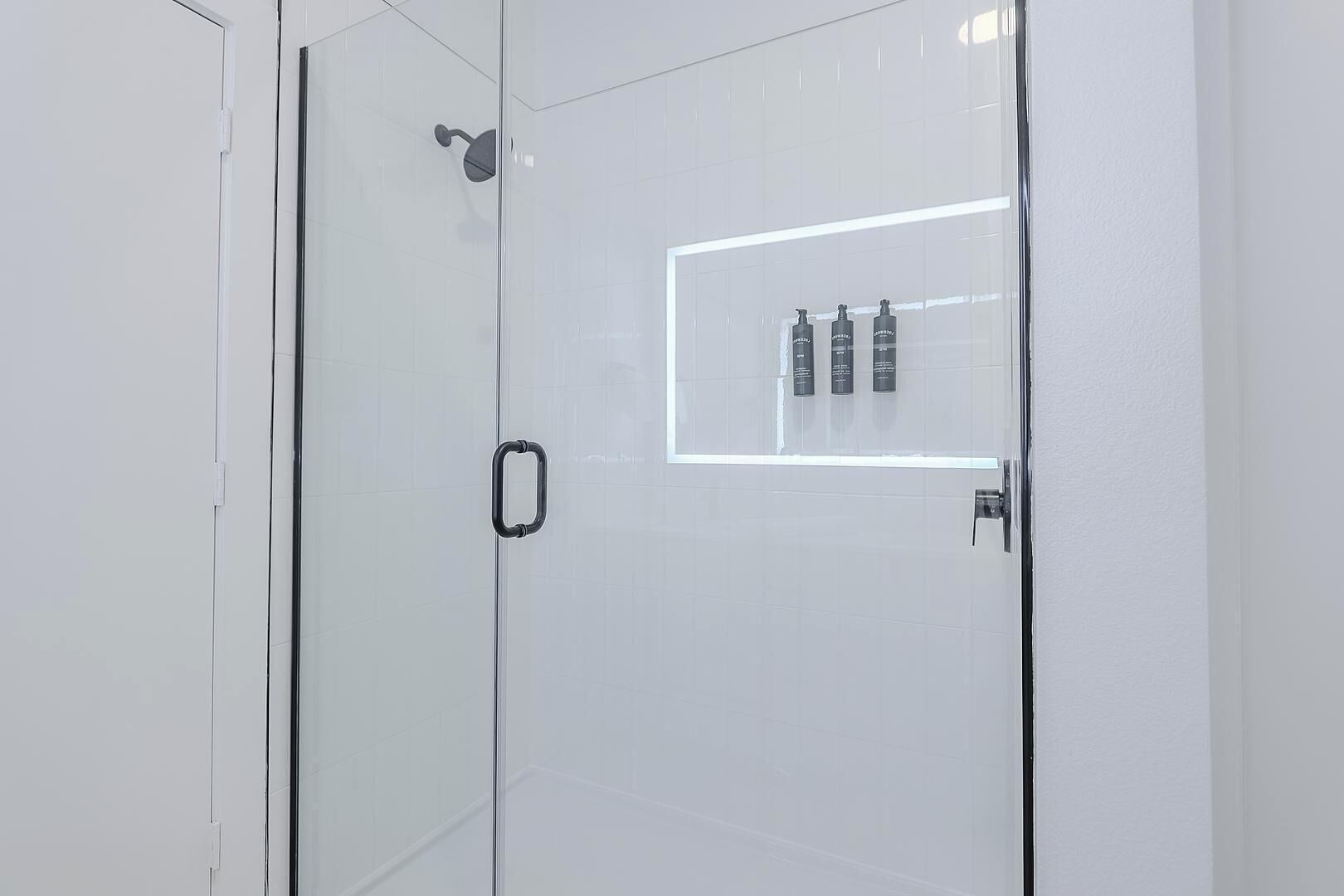 A modern bathroom with a glass shower enclosure, a visible showerhead, and a shelf with toiletries.
