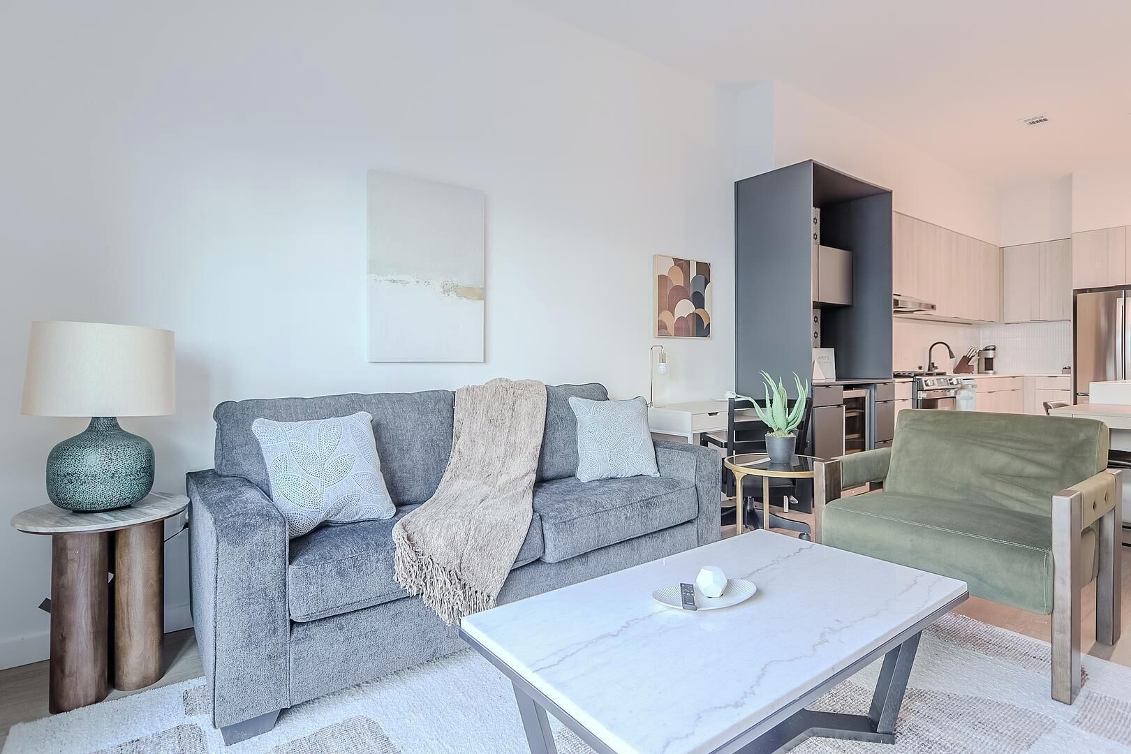 Een moderne woonkamer met een grijze bank, een salontafel, een lamp en een keuken in de achtergrond.