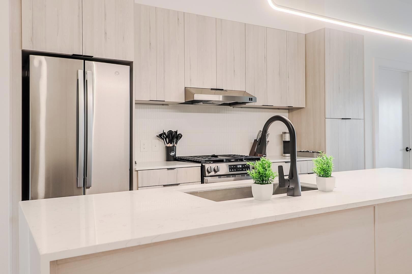 Una cocina moderna con un refrigerador de acero inoxidable, una estufa a gas, un grifo negro y dos plantas en macetas sobre la encimera.