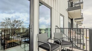A balcony with two chairs and a table, a glass door, and a view of the street and buildings. - Landing | Beautiful 2BD, Courtyard, Gym (Houston)