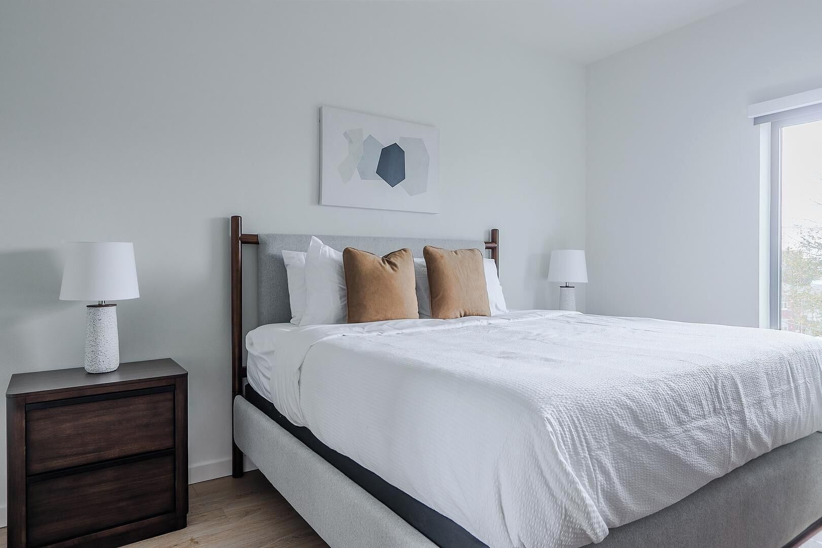 A bedroom with a bed, two bedside tables, a window, and a wall-mounted artwork.