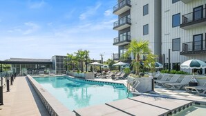 Uma piscina com espreguiçadeiras e guarda-sóis, cercada por um edifício moderno com varandas.