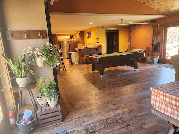 A pool table in a room with wooden flooring, a kitchen area in the background, and potted plants.
