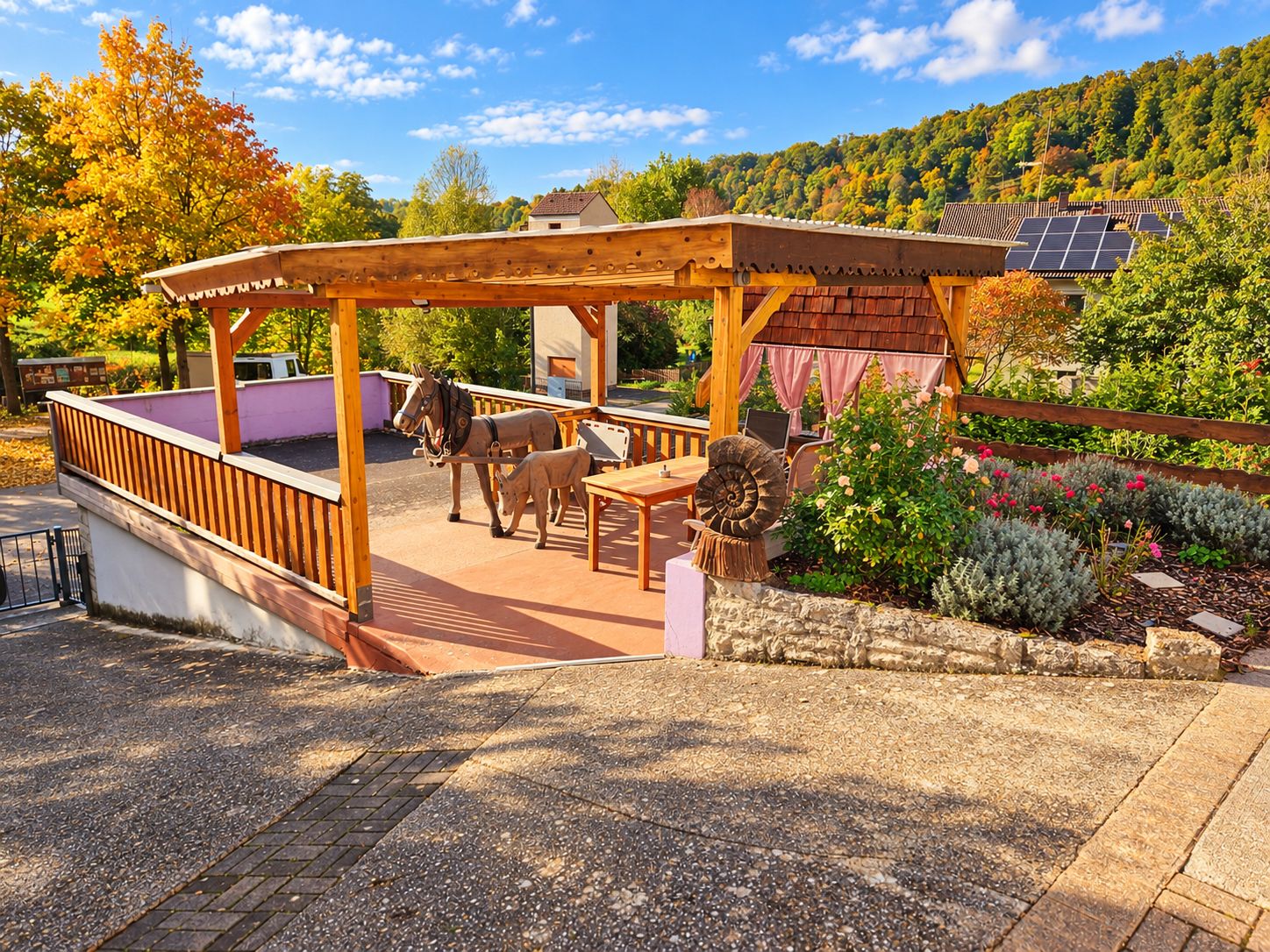 Eine hölzerne Pergola mit Sitzgelegenheiten, eine Pferdestatue und ein Garten mit verschiedenen Pflanzen.