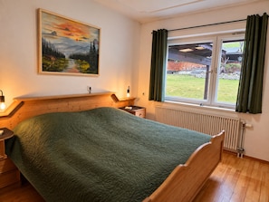 Ein Schlafzimmer mit einem hölzernen Bett, einem Nachttisch, einer Lampe, einem Gemälde, einem Fenster mit Vorhängen und einer Heizung.