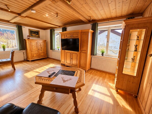 Ein Zimmer mit Holzwandverkleidung, einem Fernseher, einer Vitrine, einem Tisch mit Platzdeckchen und einem Fenster mit Blick ins Freie.