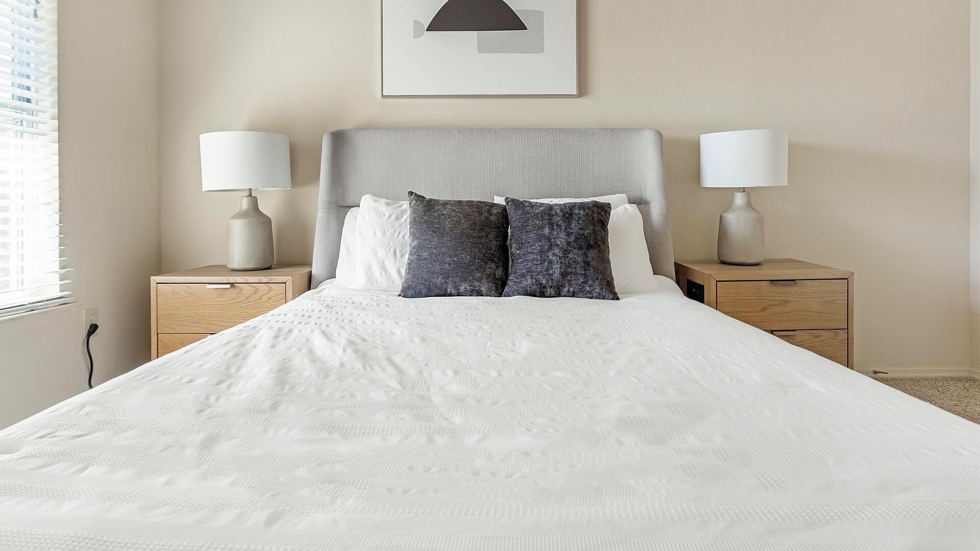 Una cama bien hecha con edredón blanco, dos mesitas de noche con lámparas, un cabecero gris y un cuadro en la pared.
