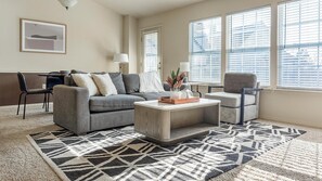 A living room with a grey sofa, a coffee table, a patterned rug, and a window with blinds.