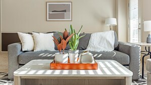 Ein Wohnzimmer mit einem grauen Sofa, weißen Kissen, einem Couchtisch mit einer Pflanzentafel und einer Lampe.