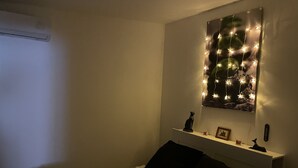 A bedroom with a bed, a white headboard, a shelf with a cat figurine, a picture frame, and a wall-mounted decoration with lights. - Apartment T3 DRC cosy climate 2 min beach (Palavas-les-Flots)