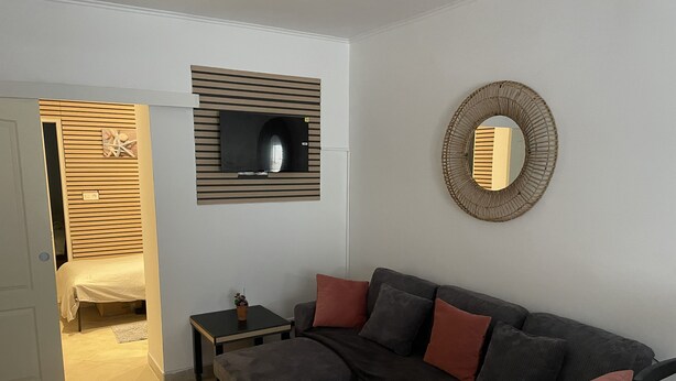 A modern living room with a sectional sofa, a small side table, a flat-screen TV mounted on a wooden panel, a round mirror on the wall, and a doorway leading to another room. - Apartment T3 DRC cosy climate 2 min beach (Palavas-les-Flots)