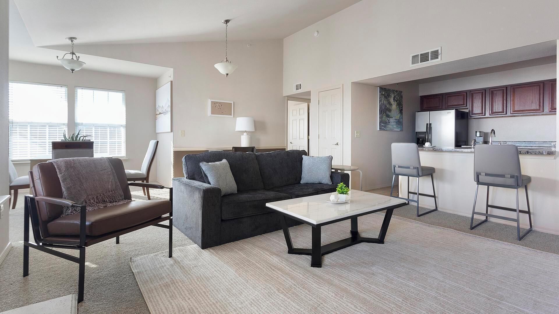 A modern living room with a sofa, chairs, a coffee table, and a kitchen area with a fridge and bar stools.
